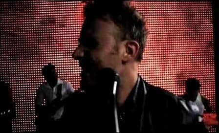 Jason Hartman singing at a microphone before a wall of red lights, a still from the music video On The Run